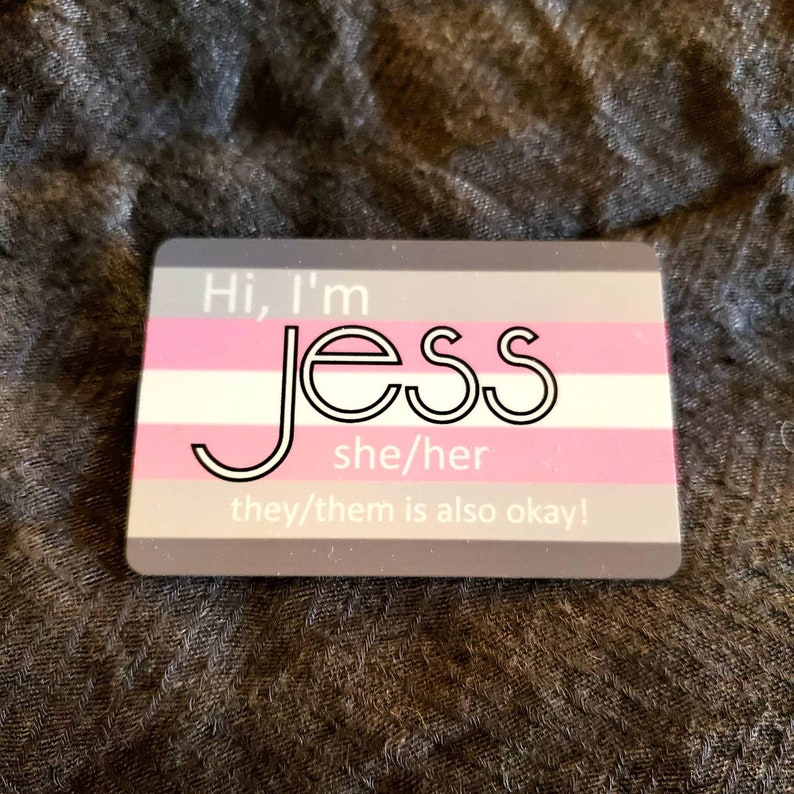 Pu&ograve; includere: Una carta grigia e rosa con il testo "Hi, I'm Jess she/her they/them is also okay!" La carta ha un design di bandiera transgender pride.