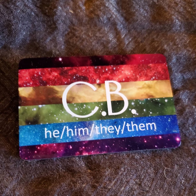 Pu&ograve; includere: Una carta color arcobaleno con uno sfondo di galassia. La carta ha le iniziali "C.B." in testo bianco e i pronomi "he/him/they/them" in testo bianco.