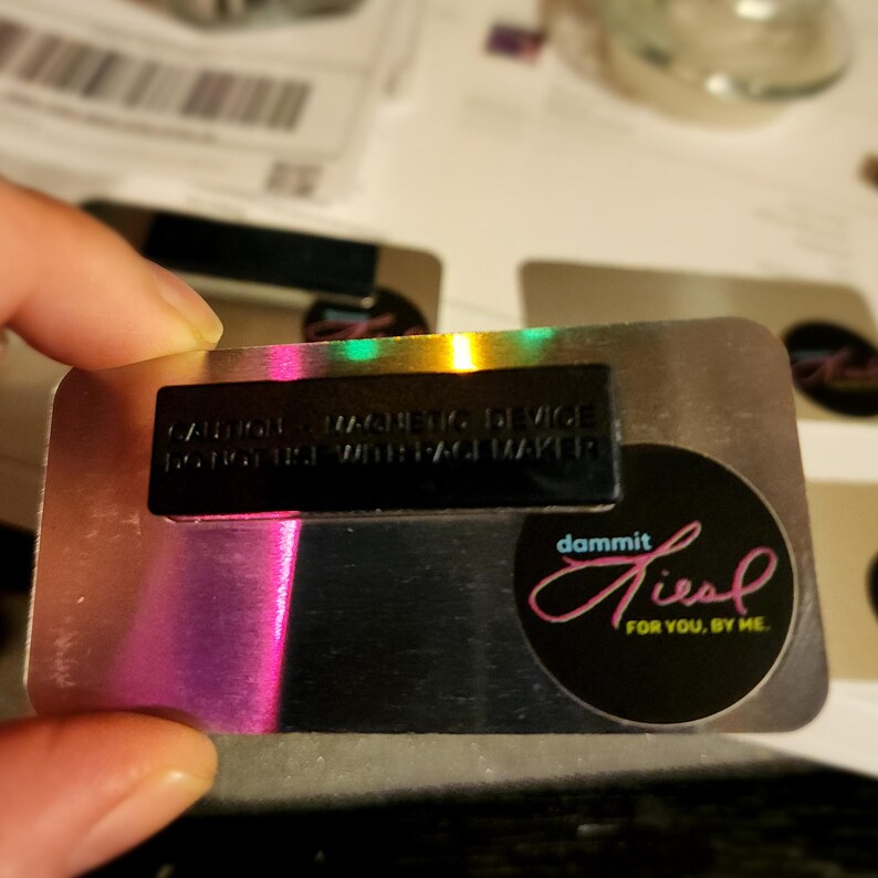 May include: A silver metal badge with a black magnetic device that says "CAUTION MAGNETIC DEVICE DO NOT USE WITH PACEMAKER". The badge also has a black circle with the text "dammit I feel for you, by me." in pink.