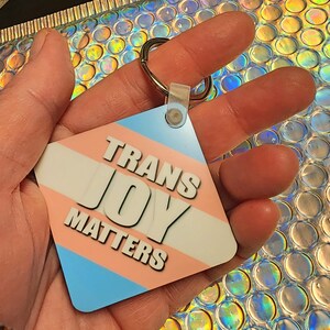 May include: A blue, pink, and white diamond-shaped keychain with the text "Trans Joy Matters".