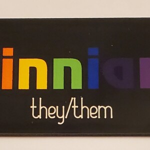 May include: Black name tag with the name "Finnian" in rainbow colours and the text "they/them" in white.