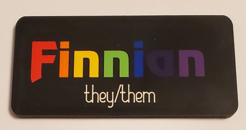 May include: Black name tag with the name "Finnian" in rainbow colours and the text "they/them" in white.