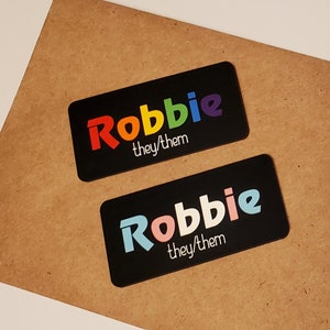 May include: Two black rectangular name tags with the name "Robbie" printed in rainbow colours. The text "they/them" is printed below the name in white.