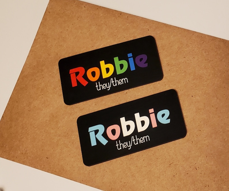 May include: Two black rectangular name tags with the name "Robbie" printed in rainbow colours. The text "they/them" is printed below the name in white.