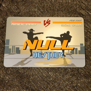 Puede incluir: Una ilustraci&oacute;n digital de un juego de lucha con dos siluetas de personas luchando. El juego se llama "NULL" y los personajes est&aacute;n etiquetados como "Energ&iacute;a f&iacute;sica" y "Cucharas sociales". El texto "HE/THEY" se muestra debajo del t&iacute;tulo del juego.