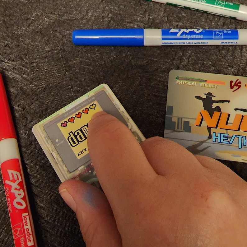 Puede incluir: Primer plano de una mano interactuando con una consola de juegos port&aacute;til Game Boy Color. La pantalla muestra un medidor de salud en forma de coraz&oacute;n pixelado con la palabra "dam" y las letras "XEY" visibles. La consola est&aacute; sobre una superficie gris.