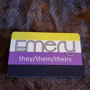 Pu&ograve; includere: Un'etichetta nominativa gialla, bianca, viola e nera con la parola "Emery" in bianco e il testo "they/them/theirs" in bianco.