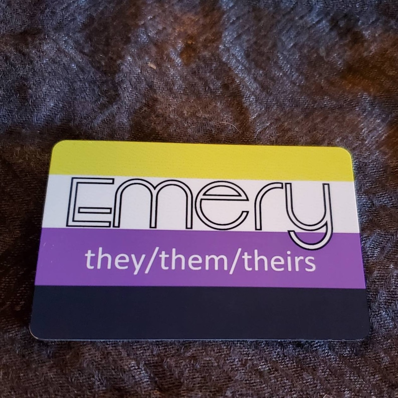 Pu&ograve; includere: Un'etichetta nominativa gialla, bianca, viola e nera con la parola "Emery" in bianco e il testo "they/them/theirs" in bianco.
