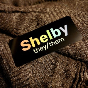 May include: A black name tag with the name "Shelby" in rainbow colours and the text "they/them" in white.