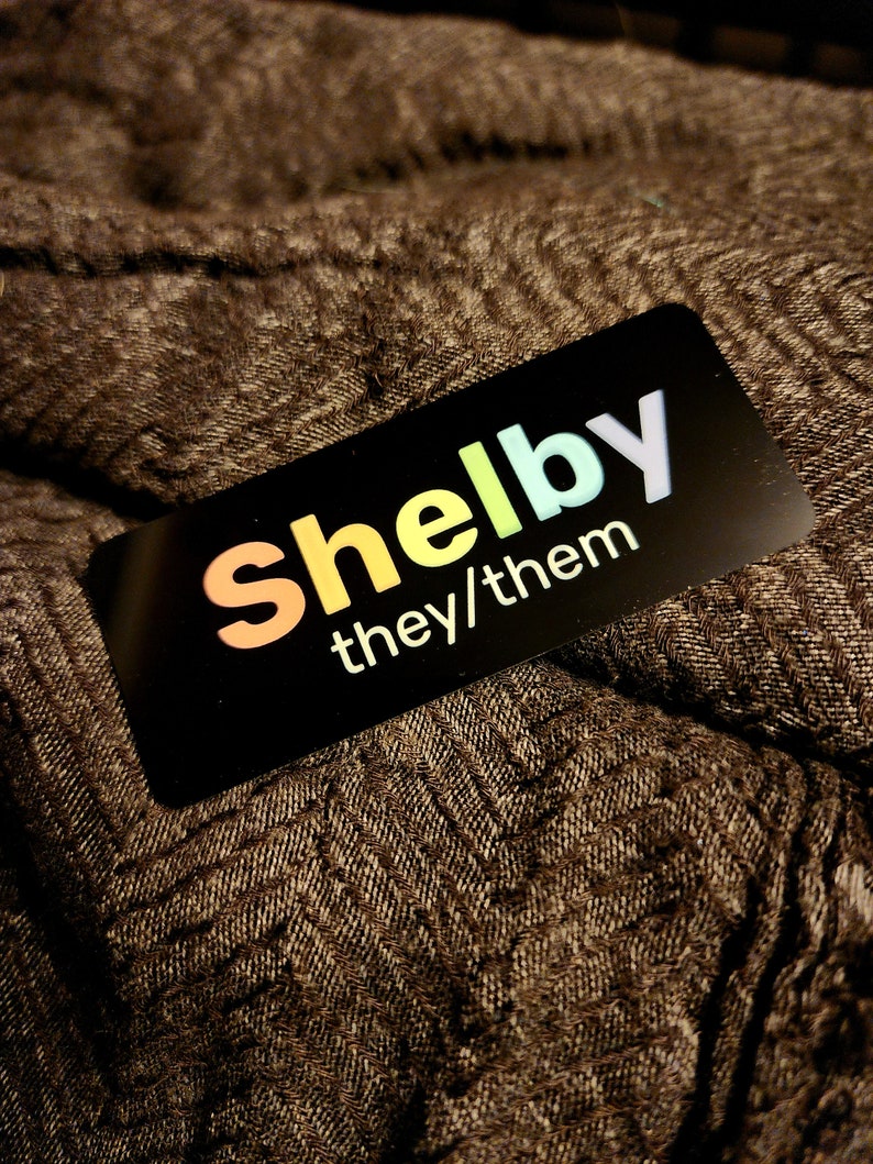 May include: A black name tag with the name "Shelby" in rainbow colours and the text "they/them" in white.