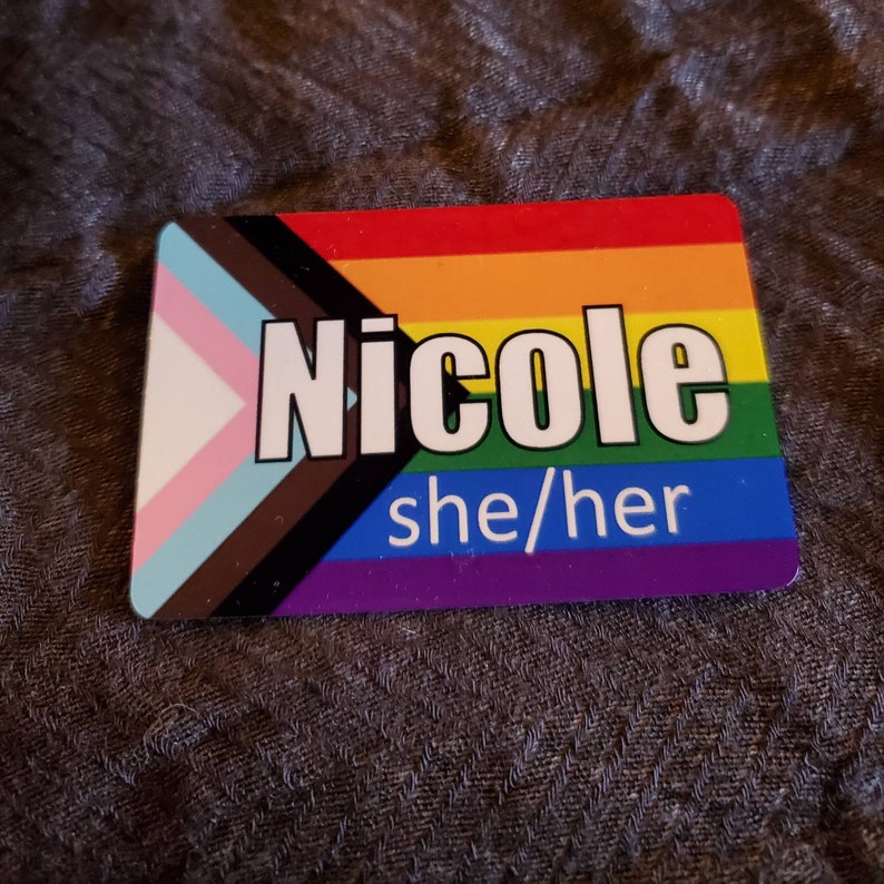 Pu&ograve; includere: Una bandiera arcobaleno del pride con una striscia nera e marrone, una bandiera del pride transgender e una striscia viola. Il nome "Nicole" &egrave; stampato in lettere bianche sulla bandiera, e il testo "lei/lei" &egrave; stampato in lettere bianche nella parte inferiore della bandiera.