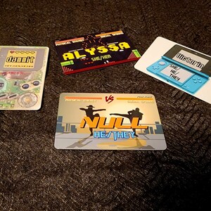 Puede incluir: Cuatro tarjetas coloridas con tem&aacute;tica de videojuegos. Las tarjetas presentan los nombres "dammit", "Alyssa", "Null" y "Victoria" con los pronombres correspondientes. Las tarjetas tienen un dise&ntilde;o pixelado y est&aacute;n colocadas sobre un fondo marr&oacute;n.