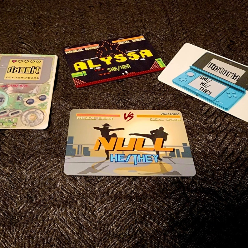 Puede incluir: Cuatro tarjetas coloridas con tem&aacute;tica de videojuegos. Las tarjetas presentan los nombres "dammit", "Alyssa", "Null" y "Victoria" con los pronombres correspondientes. Las tarjetas tienen un dise&ntilde;o pixelado y est&aacute;n colocadas sobre un fondo marr&oacute;n.