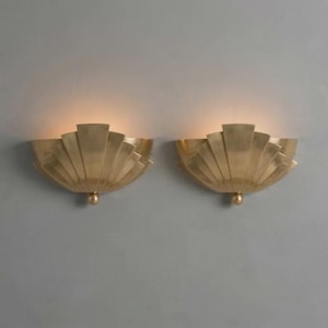 May include: Two matching wall sconces made of brushed brass metal. Each fixture features a semi-circular, fan-like shell design with radiating vertical panels and a small spherical accent at the bottom centre, emitting a soft warm glow.