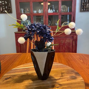 May include: A ceramic vase with a dark brown glaze and a central triangular beige textured panel. The vase holds an arrangement of dark blue chrysanthemum-like flowers and thin stems topped with small, round, white fuzzy pom-pom blossoms.