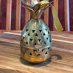 May include: A polished brass pineapple-shaped container featuring a hinged lid with a latch closure and triangular cutouts throughout the body. The top includes a decorative leaf-shaped finial and a thin, light-colored ribbon attached to the side.