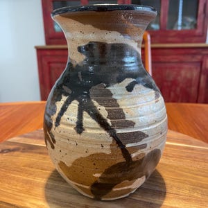 May include: A ceramic vase with a flared rim and a rounded body featuring a textured surface. The exterior displays a mottled glaze pattern in shades of cream, tan, and dark brown, with horizontal ridges encircling the midsection of the vessel.
