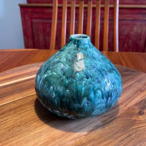 May include: A round ceramic vase featuring a glossy glaze with a blend of deep teal, forest green, and dark blue tones. The surface displays a mottled, drip-like pattern with subtle hints of metallic gold accents near the narrow circular opening.