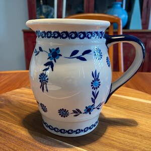 May include: A white ceramic jug featuring a hand-painted blue floral pattern with small blossoms and leaves. The jug has a curved handle, a wide rim with a pouring spout, and a decorative band of blue circular motifs around the top and base.