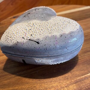 May include: A heart-shaped lidded container with a smooth, light gray base featuring black and pink paint splatters. The top surface is adorned with a textured, raised white beaded pattern that contrasts with the smooth, speckled finish below.