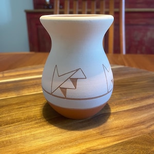 Può includere: Un piccolo vaso in ceramica arrotondato con una sezione superiore bianco opaco e una base in terracotta naturale. La parte bianca presenta motivi geometrici incisi composti da triangoli e linee che rivelano l'argilla bruno-rossastra.