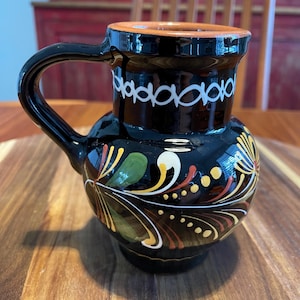 May include: A black ceramic pitcher with a glossy finish, featuring a hand-painted floral design in yellow, green, and red. The upper neck displays a white circular pattern, and the rim is a natural terracotta color with a single side handle.