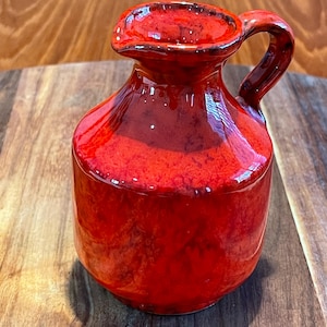 May include: A small, vibrant red ceramic pitcher with a glossy, mottled finish. The vessel features a rounded body, a short neck, a small pouring spout, and a single curved handle attached to the side, showcasing deep red and orange tones.