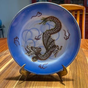 May include: A round ceramic decorative plate featuring a central illustration of a brown and white dragon with a long, winding body. The plate has a wide, gradient blue border with gold trim and small red calligraphic symbols around the dragon.