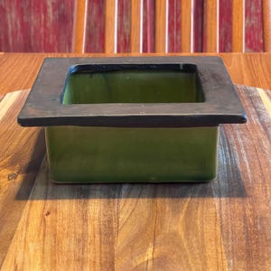 May include: A square ceramic planter featuring a glossy olive green interior and exterior body, topped with a wide, flat, matte black rim that extends slightly beyond the edges of the container, creating a distinct contrast in color and texture.