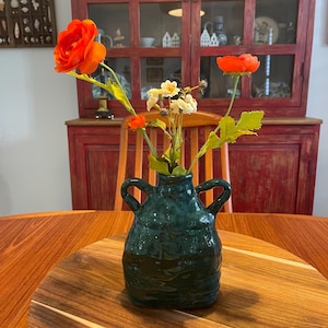 May include: A dark green ceramic vase with two side handles containing a bouquet of artificial flowers. The arrangement includes two large vibrant orange blossoms, a smaller orange bud, and a cluster of tiny white flowers with green stems.