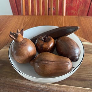 May include: A white bowl containing five carved wooden decorative fruits, including a pomegranate, an apple, a pear, a lemon, and a long, slender piece, all featuring a polished dark brown finish with visible wood grain patterns.