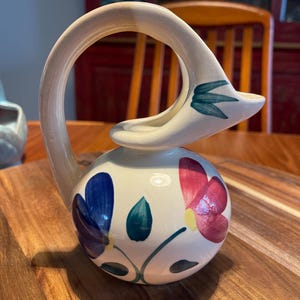 May include: A cream-colored ceramic pitcher with a rounded body and a high, arched handle. The pitcher features hand-painted floral motifs in shades of blue, pink, and green, with a stylized spout that curves upward to resemble a bird's neck.
