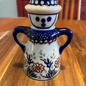 Op de afbeelding: Een keramieken beeldje met twee handvatten, voorzien van een witte basis met blauwe, bruine en groene bloemmotieven. Het figuurtje heeft een ronde blauwe hoed, een eenvoudig gezicht met drie blauwe stippen en een uitlopende voet met bloemen.