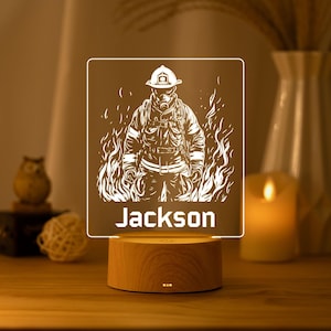Puede incluir: Una luz nocturna cuadrada de acrílico con una ilustración de líneas blancas de un bombero con equipo completo rodeado de llamas. El nombre Jackson está impreso en letras de bloque blancas debajo de la figura. La luz descansa sobre una base de madera.