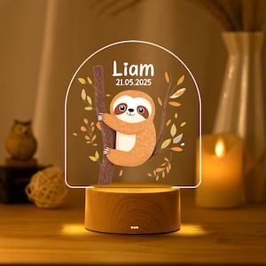 May include: An arched acrylic night light featuring a cartoon sloth clinging to a brown tree branch with orange and yellow leaves. The text "Liam" and "21.05.2025" is printed above the sloth. The light sits on a round, light-colored wooden base.