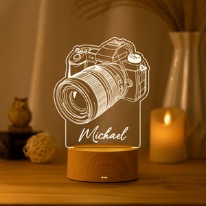 May include: A clear acrylic night light featuring a white line-art illustration of a DSLR camera with a lens and the name Michael printed in script font. The acrylic piece is mounted on a round, light-colored wooden base with a power button.