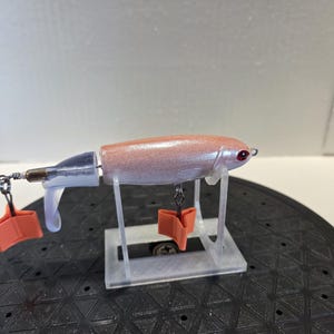 May include: A fishing lure with a shimmering light pink body, a red eye, and a translucent tail section. It features two bright orange, three-pronged hook covers attached to the metal hooks. The lure is positioned on a clear plastic display stand.