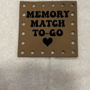 Könnte beinhalten: Ein quadratisches braunes Schild mit abgerundeten Ecken und einer Reihe kleiner, kreisförmiger Löcher entlang des Randes. Der Text "MEMORY MATCH TO-GO" ist in schwarzer, fetter, abgerundeter Schrift gedruckt, gefolgt von einem schwarzen Herz.