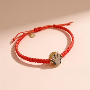 Puede incluir: Una pulsera de cordón trenzado rojo con un dije dorado en forma de concha. El dije está adornado con piedras blancas transparentes y pequeñas piedras azul oscuro dispuestas en forma de abanico. La pulsera incluye un cierre de nudo corredizo.