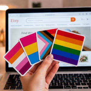 Könnte beinhalten: Vier quadratische Aufkleber mit verschiedenen Pride-Flaggen-Designs, darunter die lesbische Flagge, die pansexuelle Flagge, die Progress-Pride-Flagge und die klassische sechsstreifige Regenbogenflagge, fächerförmig vor einem Etsy-Laptopbildschirm.