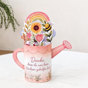 May include: A pink watering can-shaped pop-up card featuring a rainbow, sunflower, tulip, and bee illustration. The text reads, "Danke, dass du uns beim Wachsen geholfen hast." The design includes a handle, spout, and floral accents at the base.