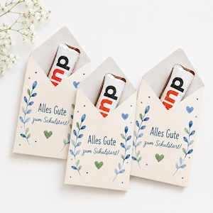 May include: Three cream-colored paper envelopes, each containing a chocolate bar. The envelopes feature blue and green watercolor leaf illustrations, small hearts, and the printed text "Alles Gute zum Schulstart!" in a dark blue script font.