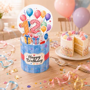 May include: A cylindrical pop-up birthday card featuring the number 12, colorful balloons, and gift boxes. The base is blue with vertical stripes and a label that reads Happy Birthday. The card is displayed on a wooden surface with confetti.