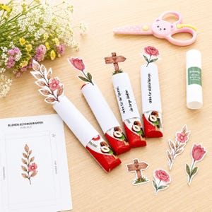 May include: Four chocolate bars wrapped in white paper sleeves with floral and wooden sign toppers. The sleeves feature German text. Nearby are loose paper cutouts of flowers and signs, a glue stick, and pink bear-shaped scissors.