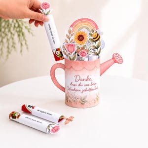 May include: A pink paper watering can centrepiece featuring a floral arrangement with a sunflower, pink flowers, a rainbow, and a bee. The text reads, "Danke, dass du uns beim Wachsen geholfen hast." Includes small paper tubes labelled "Danke f&uuml;r starke Nerven."