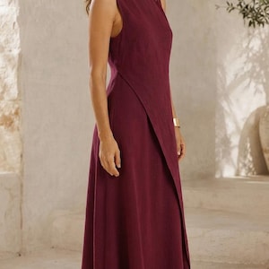 May include: A sleeveless, floor-length dress in a deep burgundy color. The garment features a high, round neckline and a distinctive wrap-style front panel that creates an asymmetrical hemline. The fabric has a visible, slightly textured weave.