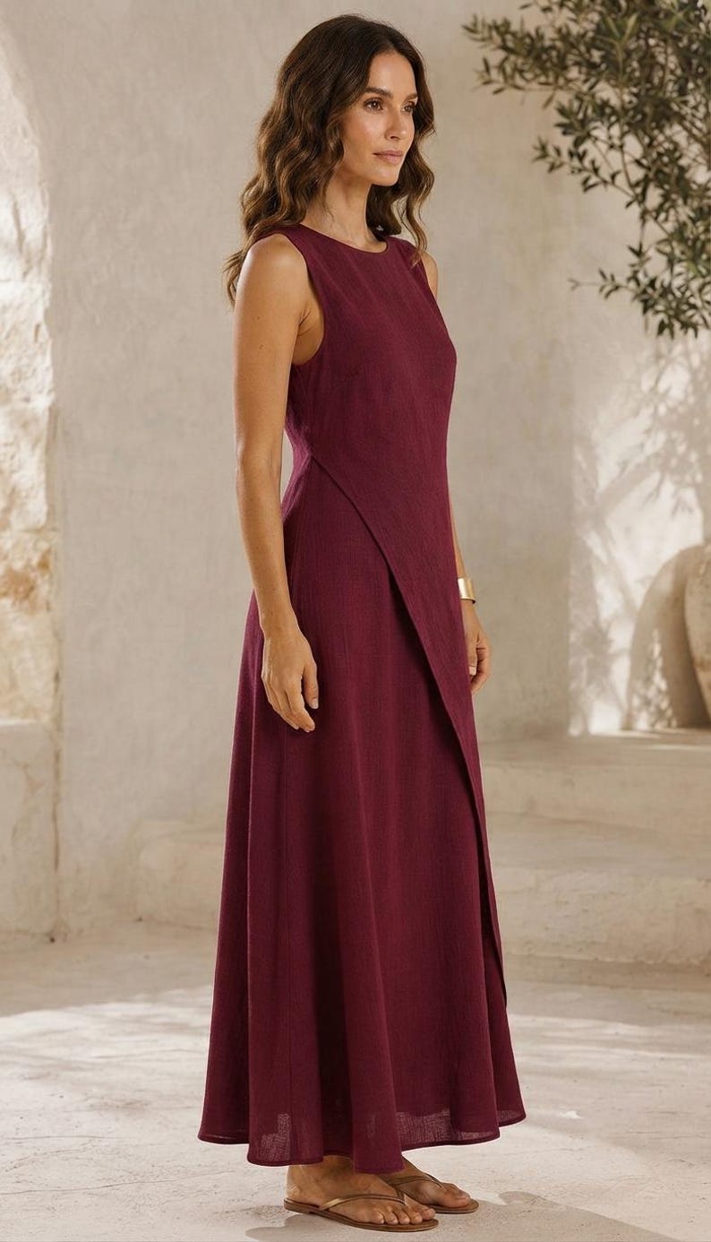 May include: A sleeveless, floor-length dress in a deep burgundy color. The garment features a high, round neckline and a distinctive wrap-style front panel that creates an asymmetrical hemline. The fabric has a visible, slightly textured weave.
