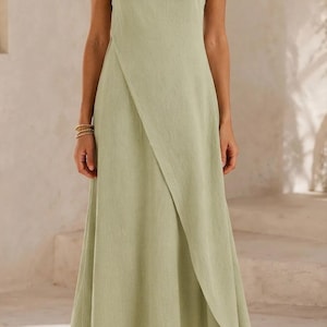 May include: A sleeveless sage green maxi dress featuring a round neckline and a distinctive overlapping front panel design that creates a diagonal hemline. The garment has a fitted bodice that transitions into a flowing, floor-length skirt silhouette.