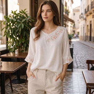 Könnte beinhalten: Eine weiße Bluse mit V-Ausschnitt und Dreiviertelärmeln mit ausgestellten Bündchen. Das Kleidungsstück ist mit zarten Blumenstickereien in Rosa- und Grüntönen an der linken Schulter sowie an den Ärmelbündchen verziert.