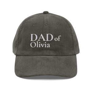 Könnte beinhalten: Eine dunkelgraue Baseballkappe aus Kord mit dem weiß gestickten Text DAD of Olivia mittig auf der Vorderseite. Die Kappe hat eine strukturierte Krone, einen gebogenen Schirm und einen Knopf an der Oberseite aus strukturiertem Stoff.